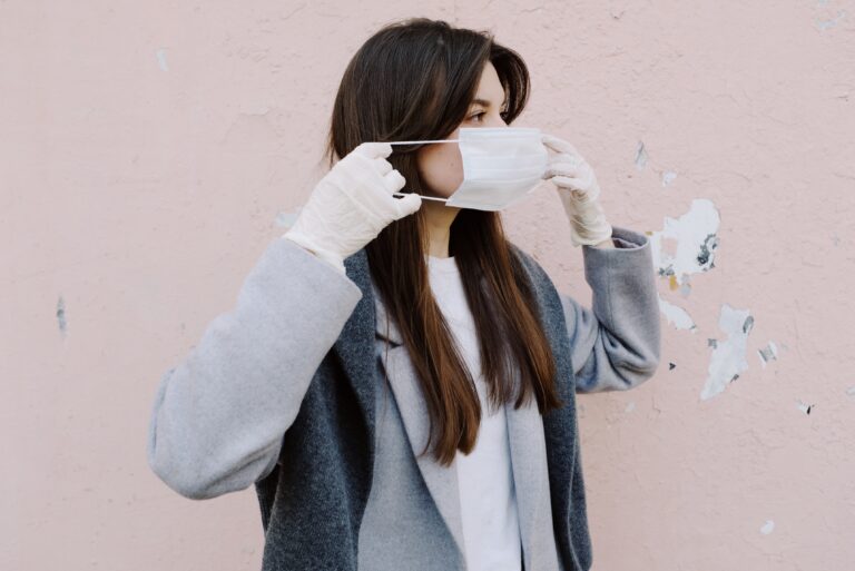 woman with long brown hair putting on a mask with gloves in front of a pink wall
