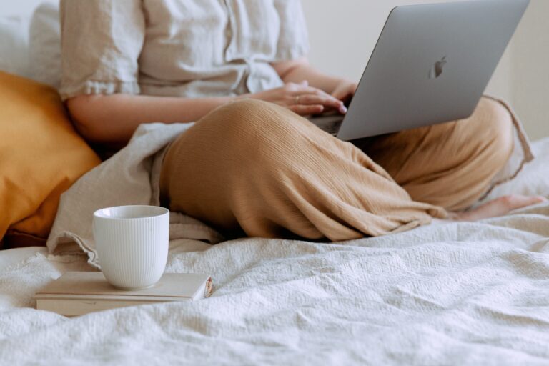 woman on her macbook on her bed with a cup of coffee and a book