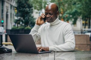 a black man in a white turtleneck on the phone and on his computer.